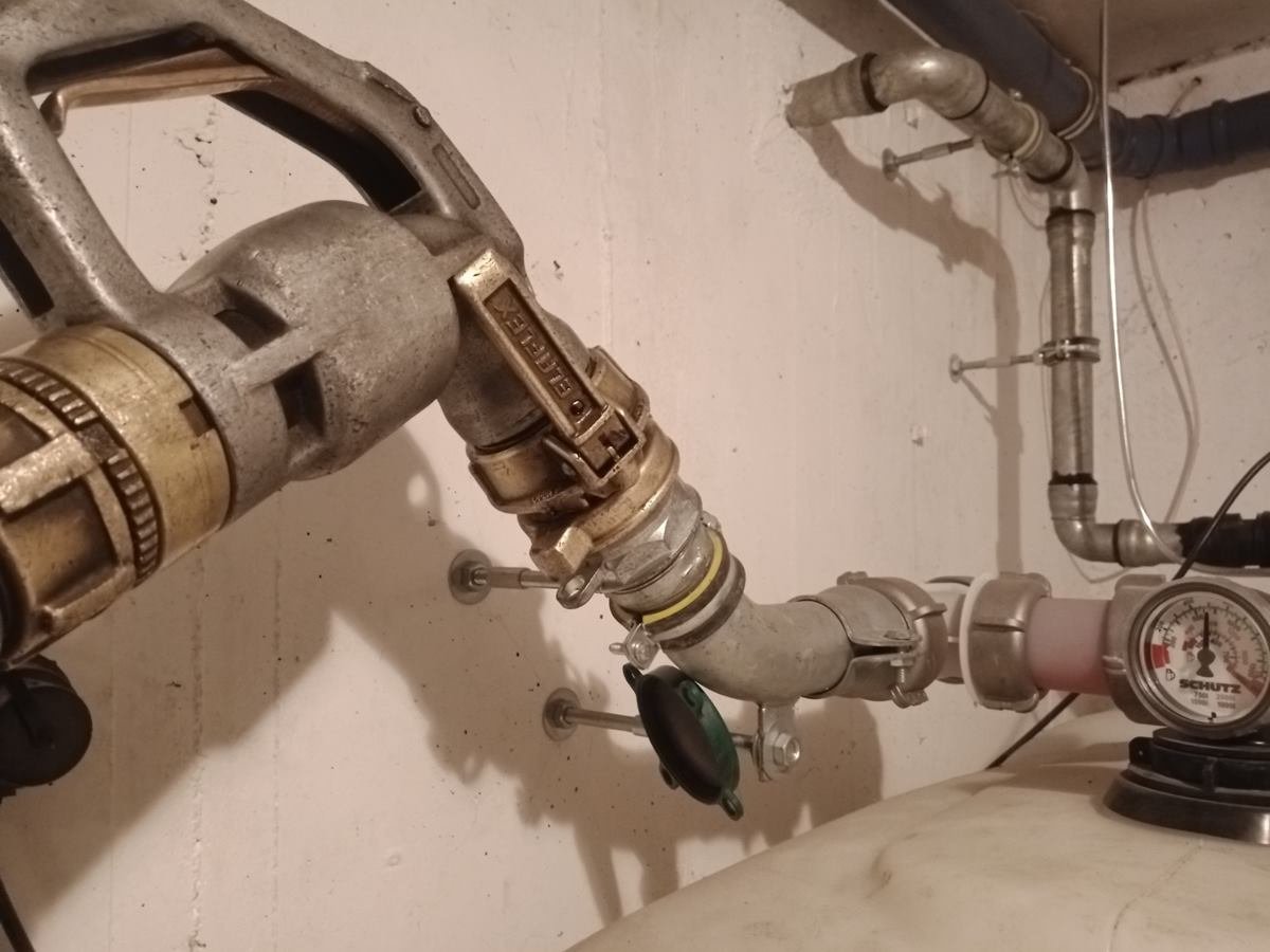 Zapfventil des Füllschlauchs, angekuppelt an den Einfüllstutzen eines Heizöltanks im Keller, rechts daneben die Füllstandsanzeige am Tank