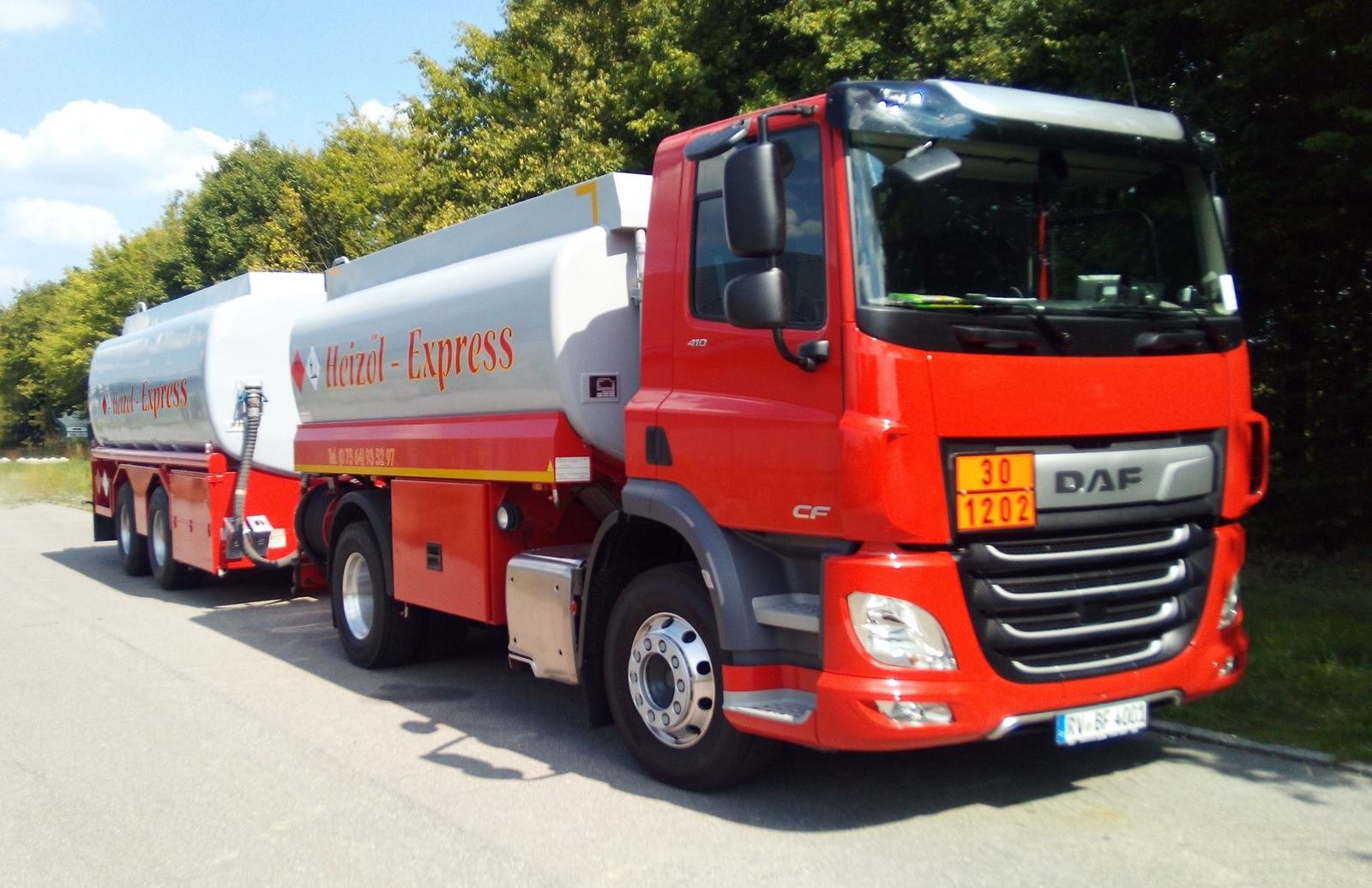 Roter Tankwagen von Heizöl-Express mit silbernem Tank und Anhänger, geparkt an einer Straße vor hohen grünen Bäumen
